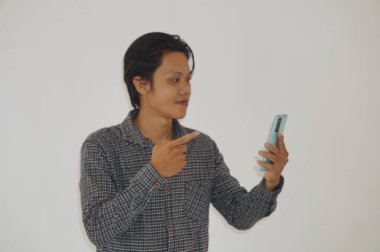 Siyah tişörtlü genç Asyalı adam şaşırdı ve beyaz arka planda izole akıllı telefona işaret etti.