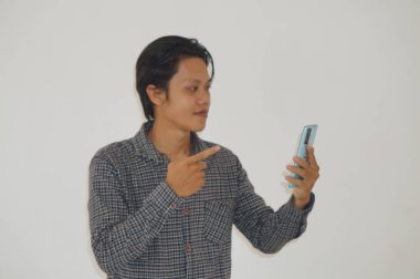 Siyah tişörtlü genç Asyalı adam şaşırdı ve beyaz arka planda izole akıllı telefona işaret etti.