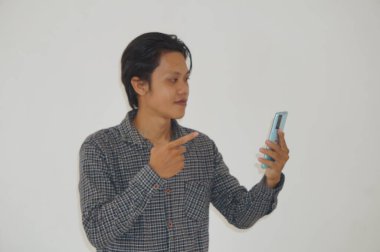 Siyah tişörtlü genç Asyalı adam şaşırdı ve beyaz arka planda izole akıllı telefona işaret etti.