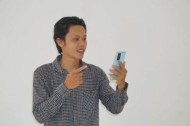 Siyah tişörtlü genç Asyalı adam şaşırdı ve beyaz arka planda izole akıllı telefona işaret etti.