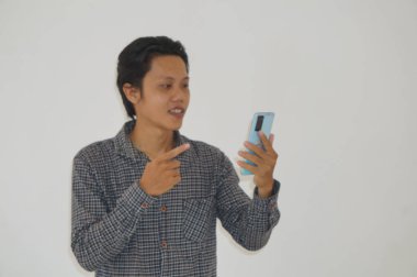 Siyah tişörtlü genç Asyalı adam şaşırdı ve beyaz arka planda izole akıllı telefona işaret etti.