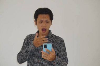 Siyah tişörtlü genç Asyalı adam beyaz arka planda izole edilmiş akıllı telefona bakarken şaşırdı.