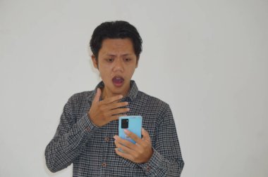 Siyah tişörtlü genç Asyalı adam beyaz arka planda izole edilmiş akıllı telefona bakarken şaşırdı.