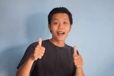 Asyalı adam kameraya gülümsüyor. Siyah tişört giyiyor. Baş parmağını gösteriyor.