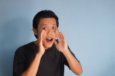 Asyalı bir adam siyah bir tişört giyiyor, ayakta duruyor ve korkmuş görünüyor. Bir eli ağzının yanında. 