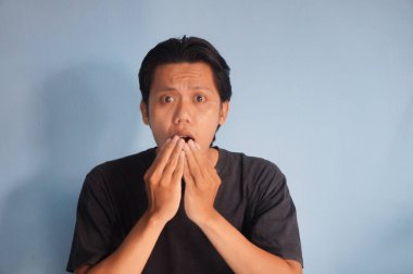 Asyalı bir adam siyah bir tişört giyiyor, ayakta duruyor ve korkmuş görünüyor. Bir eli ağzının yanında. 