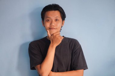 Genç Asyalı adam gülümsüyor siyah gömlekli kameraya bakıyor, el hareketleriyle çenesini tutuyor.