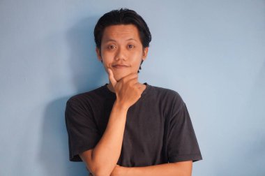 Genç Asyalı adam gülümsüyor siyah gömlekli kameraya bakıyor, el hareketleriyle çenesini tutuyor.