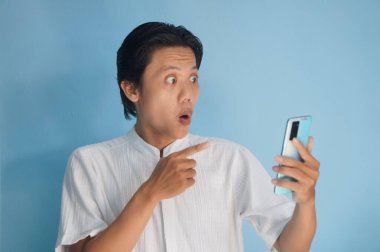 Beyaz tişörtlü genç Asyalı adam şaşırmış ve akıllı telefonu işaret ediyor. 