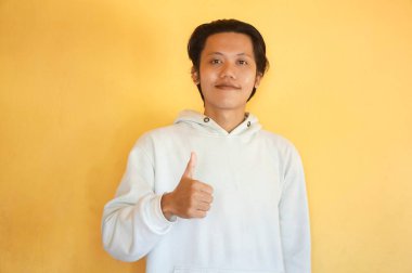 Asyalı genç adam kameraya gülümsüyor. Kapüşonlu başparmak gösteriyor.