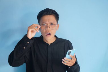 Siyah tişörtlü genç Asyalı adam şok olmuş bir ifadeyle gözlük çerçevesine dokunuyor.