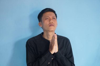 Yakışıklı Asyalı genç adam siyah tişört giyip ellerini kavuşturup af diliyor.