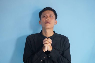 Yakışıklı Asyalı genç adam siyah tişört giyip ellerini kavuşturup af diliyor.