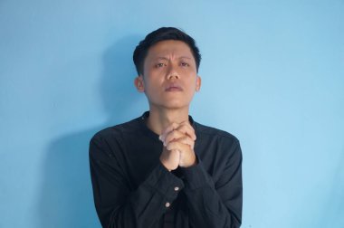 Yakışıklı Asyalı genç adam siyah tişört giyip ellerini kavuşturup af diliyor.