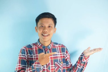 Kareli gömlek giyen genç bir Asyalı parmağını yana uzatarak gülümsüyor.