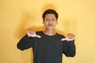 Siyah tişört giyen Asyalı genç adam tasvip etmiyormuş gibi baş parmağıyla poz veriyor.