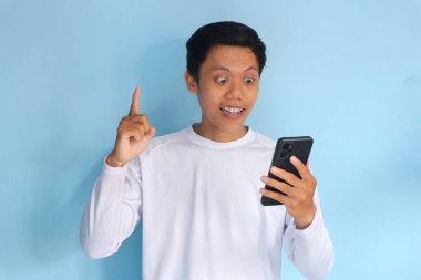 Asyalı genç adam cep telefonuna bakıp parmağını kaldırırken hayret verici bir ifadeyle bakıyor.