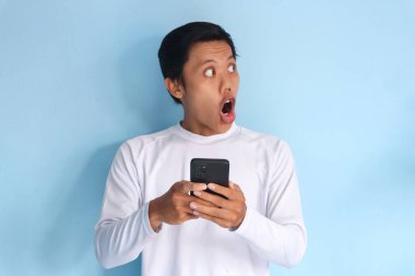 Beyaz tişört giyen Asyalı genç adam cep telefonu tutarken şok edici bir ifade sergiliyor.