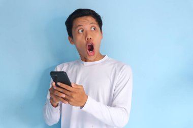Beyaz tişört giyen Asyalı genç adam cep telefonu tutarken şok edici bir ifade sergiliyor.