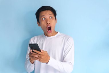 Beyaz tişört giyen Asyalı genç adam cep telefonu tutarken şok edici bir ifade sergiliyor.
