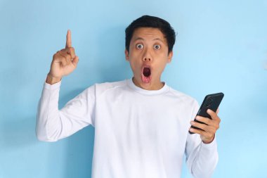 Beyaz tişört giyen Asyalı genç adam cep telefonu tutarken şok edici bir ifade sergiliyor.