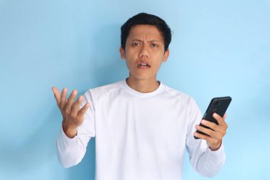 Beyaz tişört giyen Asyalı genç adam cep telefonu tutarken şaşkın bir ifade sergiliyor.