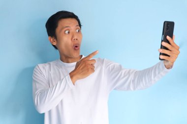 Beyaz tişört giyen Asyalı genç adam elinde cep telefonu tutarken şaşırdı.