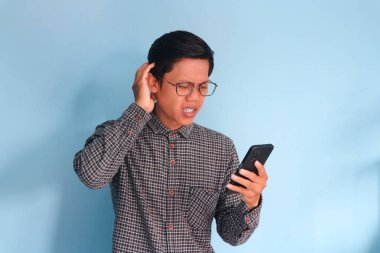 Genç Asyalı adam cep telefonunu karışık bir ifadeyle tutuyor.