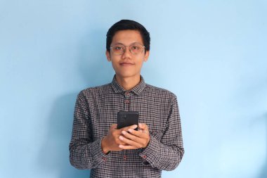 Genç Asyalı adam cep telefonu tutarken kameraya bakıp gülümsüyor.