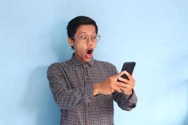 Genç Asyalı adam şok olmuş bir ifadeyle cep telefonuna bakıyor.