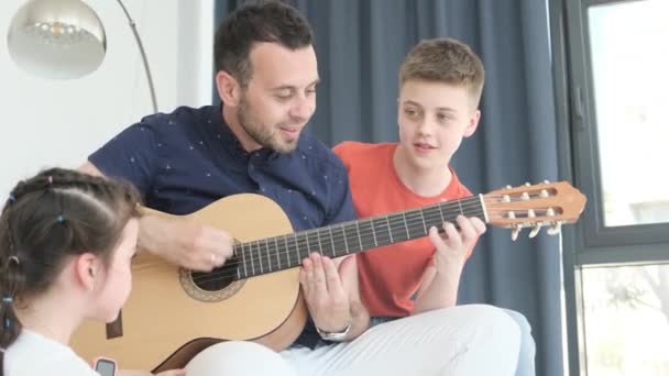 一个年轻的爸爸为他的孩子们弹奏吉他 他给孩子们表演 欢乐的家庭在家里沙发上休息 家庭幸福的概念 — 图库视频影像