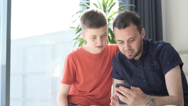 爸爸和儿子在看智能手机屏幕 他们坐在沙发上休息 父子关系 — 图库视频影像
