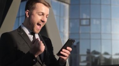 Takım elbiseli genç bir adam akıllı bir telefon tutuyor ve borsadan elde ettiği kârla seviniyor. Bir iş adamının neşesi. Adam büyük ikramiyeyi kazandı.