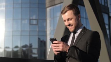 Avrupalı güler yüzlü iş adamı elinde akıllı telefon, takım elbiseli ofis çalışanı şehir sokaklarında SMS okuyor. Yavaş çekim