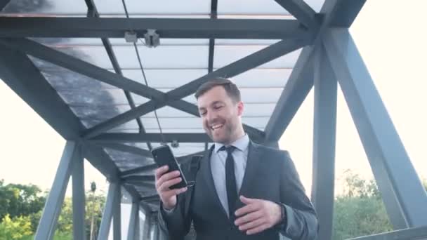 アメリカのビジネスマンはビジネスセンターの近くの屋外でスマートフォンを使ってビデオチャットをしています ビジネススーツのビジネスマンがビデオ通話で話してる — ストック動画