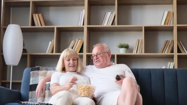 いくつかの年金受給者はソファに座っている 彼らはポップコーンを食べているとテレビを見ている 幸せな高齢者のカップル休んで自宅 — ストック動画