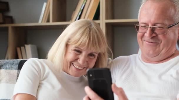 スマートフォンを持っている幸せな高齢者のカップルは 楽しそうに笑い モバイルアプリ技術を使って楽しみ 陽気なおじいちゃんたちはソファで自宅でリラックスし インターネットを使って — ストック動画