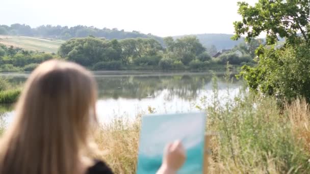 在一个温暖的夏日里 一位美丽的金发女子在湖边画了一幅自然的图画 在自然界中创造一个抽象的画面 艺术作为一种业余爱好 — 图库视频影像