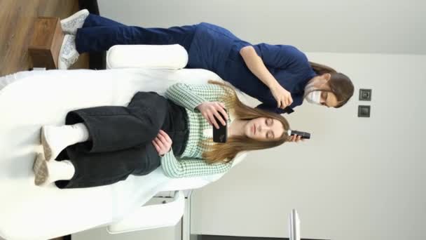 垂直ビデオでは 皮膚鏡で若い女性の頭を検査します クリニックのクライアントの手の中に皮膚や髪の可視化とスマートフォン 4Kビデオ — ストック動画