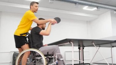 Sınırlı fiziksel yetenekleri olan genç bir adam masa tenisi oynamak için pratik yapıyor. Koçun yardımıyla sporcunun rehabilitasyonu