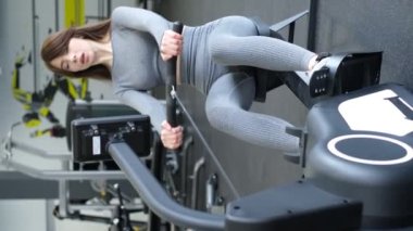 Sportif kız spor salonunda kürek egzersizi yapıyor. Gri spor kıyafetleri içinde mükemmel bir vücut yapısı olan bir kız. Dikey video
