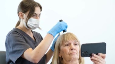 Bir kadın trikoloğu, modern bir klinikte müşterisiyle çalışıyor. Dermatoskop kullanarak kafa derisi temizleme. Yaşlı kadın saç tedavisi görüyor.