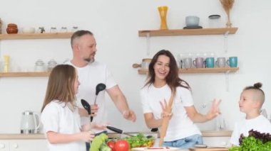 Pozitif anne, baba ve beyaz tişörtlü iki çocuk mutfakta yemek pişirirken dans ediyorlar. Mutlu aile birlikte.