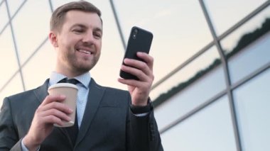 Başarılı sakallı iş adamının portresi, yönetmen, antrenör kahve içen ve akıllı telefona bakan. Modern bir gökdelenin arka planında yakışıklı bir adam..