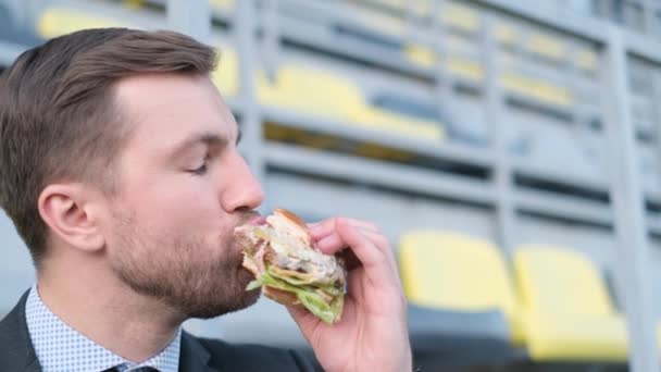 若い魅力的なビジネスマン マネージャースポーツアリーナに座ってホットドッグを食べる ファーストフードだ 有害な食べ物だ サラリーマンランチ — ストック動画