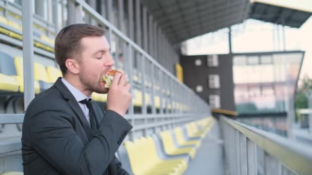 若い魅力的なビジネスマン マネージャースポーツアリーナに座ってホットドッグを食べる ファーストフードだ 有害な食べ物だ サラリーマンランチ — ストック動画