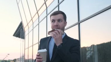 Yakışıklı sakallı adam, ofis çalışanı, yönetici, büyük pencereli bir gökdelenin arka planında duruyor. Ofis çalışanları öğle yemeği. Bir şeyler atıştır.