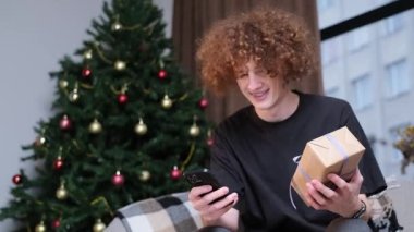 Kıvırcık saçlı, gülümseyen genç bir adam elinde yeni yıl hediyesi tutuyor ve fotoğrafını çekiyor. Sevilen bir kıza hediye. Mutlu duygular