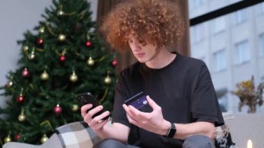 Genç kıvırcık adam akıllı telefondan alışveriş yapıyor. Kanepede oturan bir adam online bir mağazadan alışveriş yapmak için bir banka kartı kullanır. 4k video