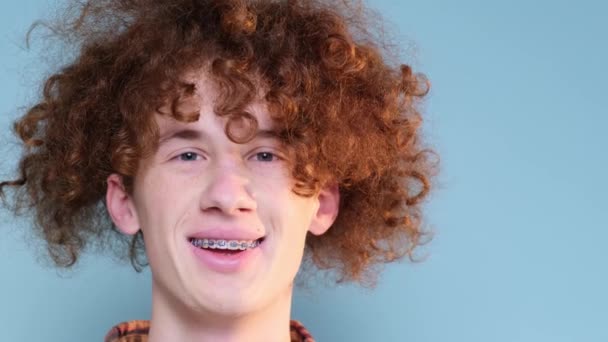 一个笑着的红头发卷发少年男孩的画像 他的牙齿上戴着牙套 背景是蓝色的工作室肖像 4K视频 — 图库视频影像